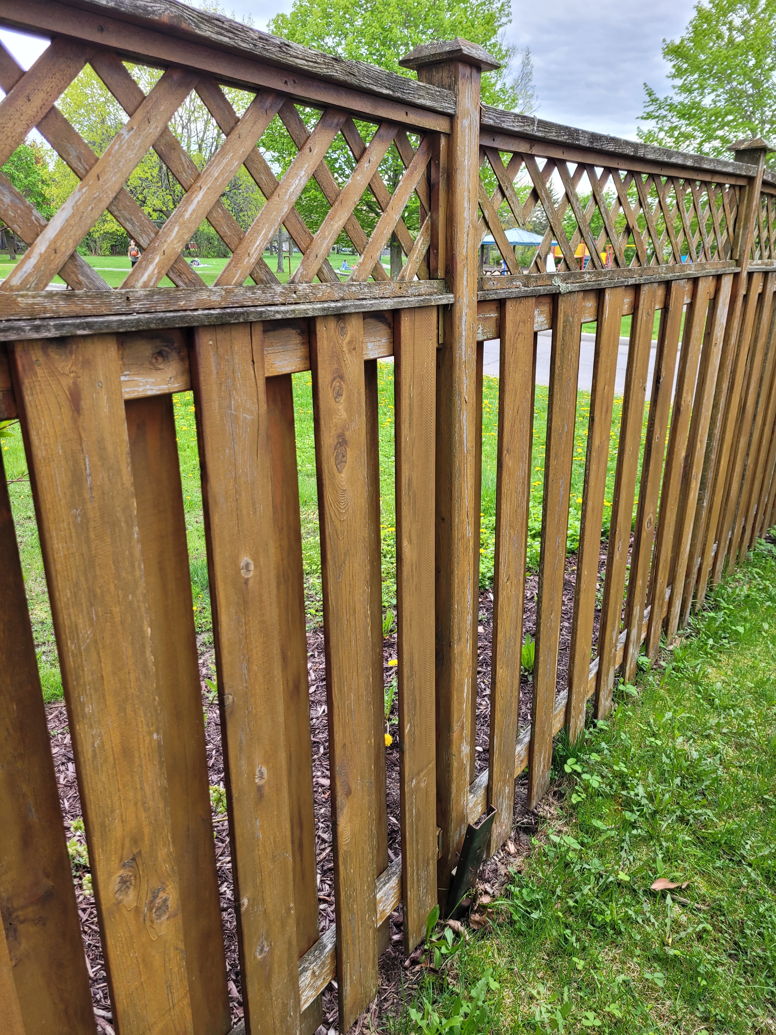 Looking down a leaning fence line to a black E-Z Mender bent over at the post base — the plate folded as the whole run tipped — Orléans, Ontario, May 2024.