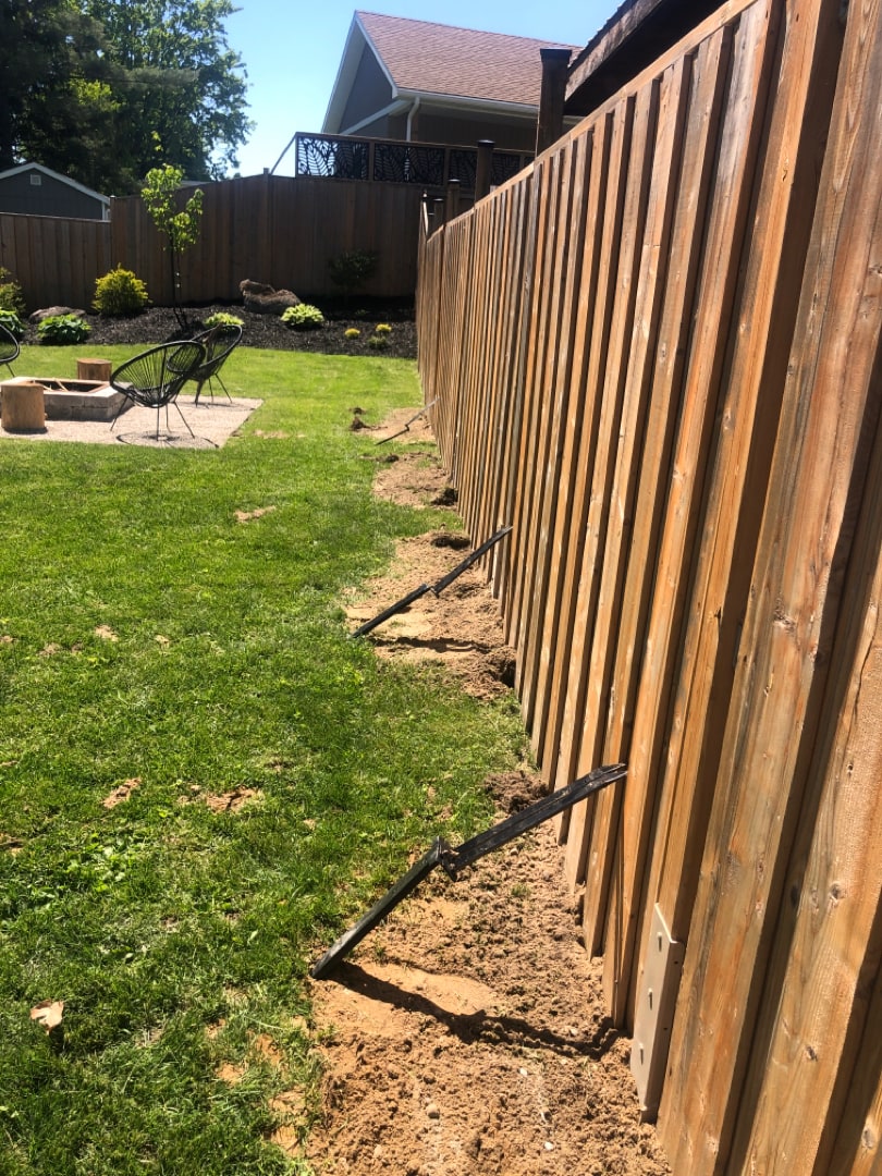 Mid-repair: failed drive-in stakes extracted and lying along the fence line while a Lean on Me structural fixture goes onto the post behind them — Kitchener, Ontario, May 2025.