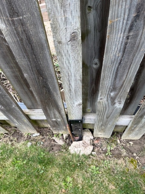 A black surface-mount post bracket from a previous repair attempt at the base of a sagging board fence, the concrete under it heaved and exposed — Mississauga, Ontario, April 2025.