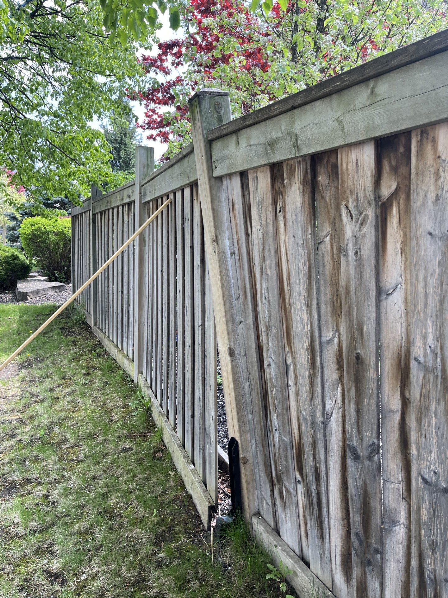 A severely leaning 6x6 fence post with a black mender plate at its base and a long lumber brace wedged against it — the emergency prop arrived after the mender failed — Whitby, Ontario, June 2025.