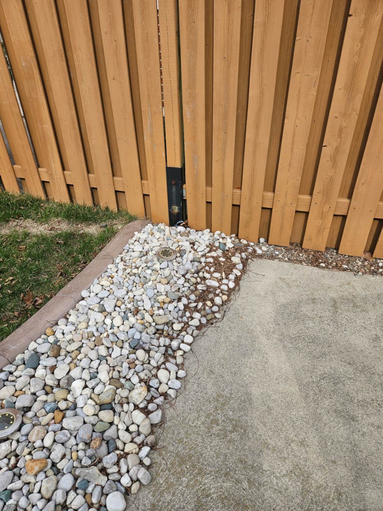An E-Z Mender bolted to the post of a well-kept, freshly stained shadowbox fence with river-rock landscaping — the post seam is splitting open directly above the plate — Windsor, Ontario, March 2026.