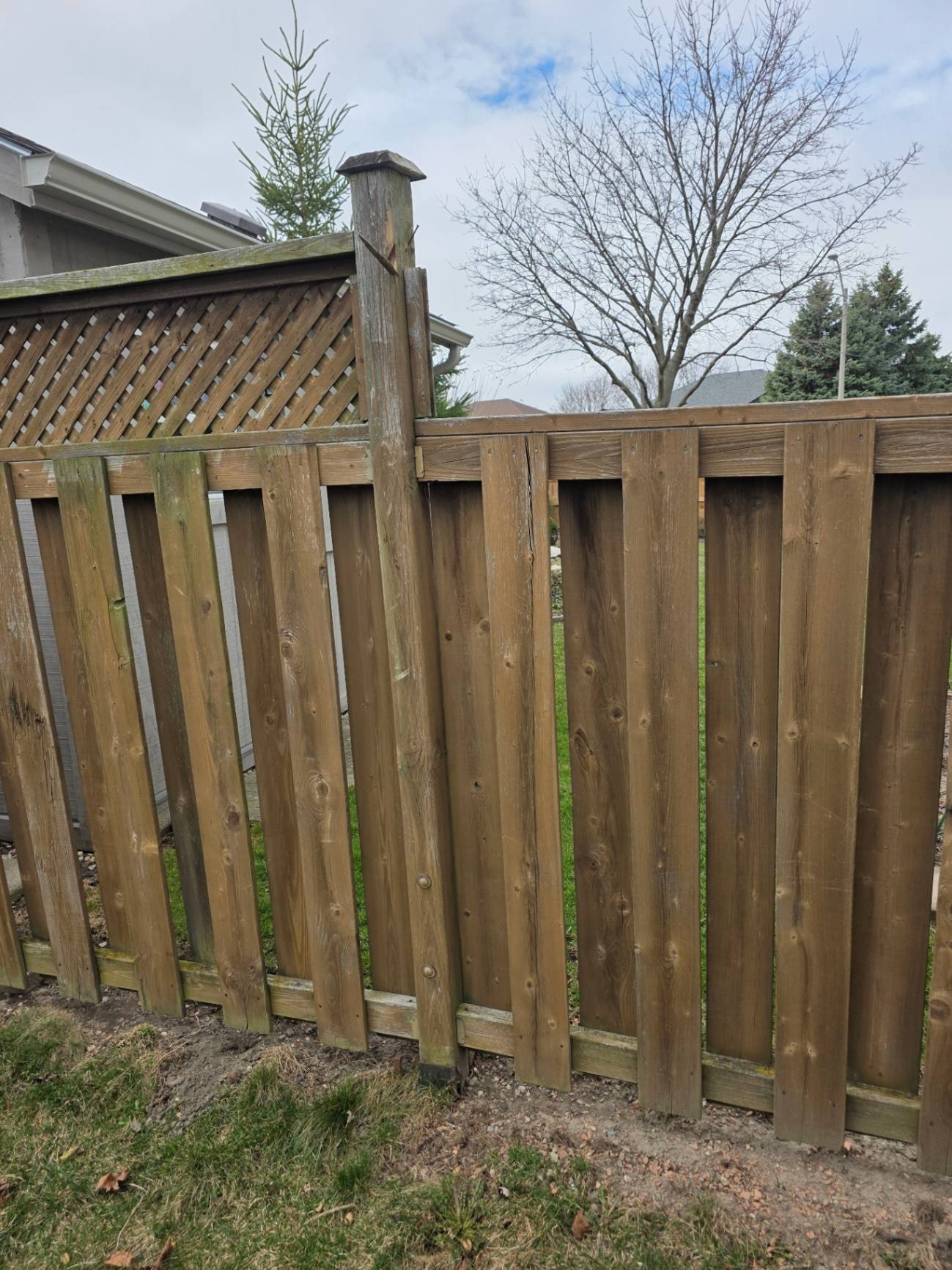 A spiked post base at the bottom of a leaning lattice-top fence post — the socket grips its few inches of wood while the post pivots freely above it — Belle River, Ontario, April 2026.