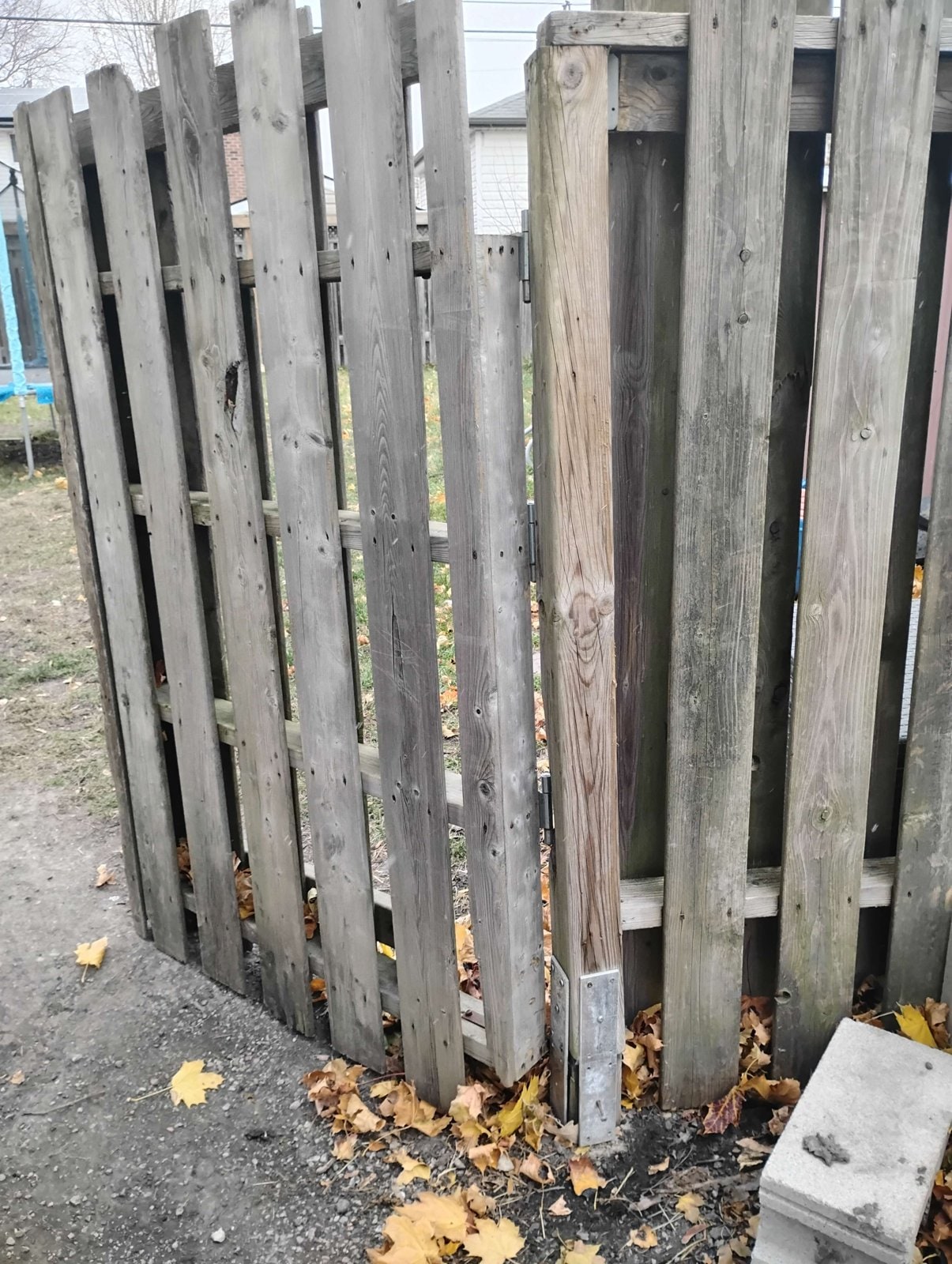 A gate post standing in a galvanized surface-mount bracket while the entire gate leans away — the bracket holds its few inches of wood and the post pivots on top of it — Windsor, Ontario, December 2025.