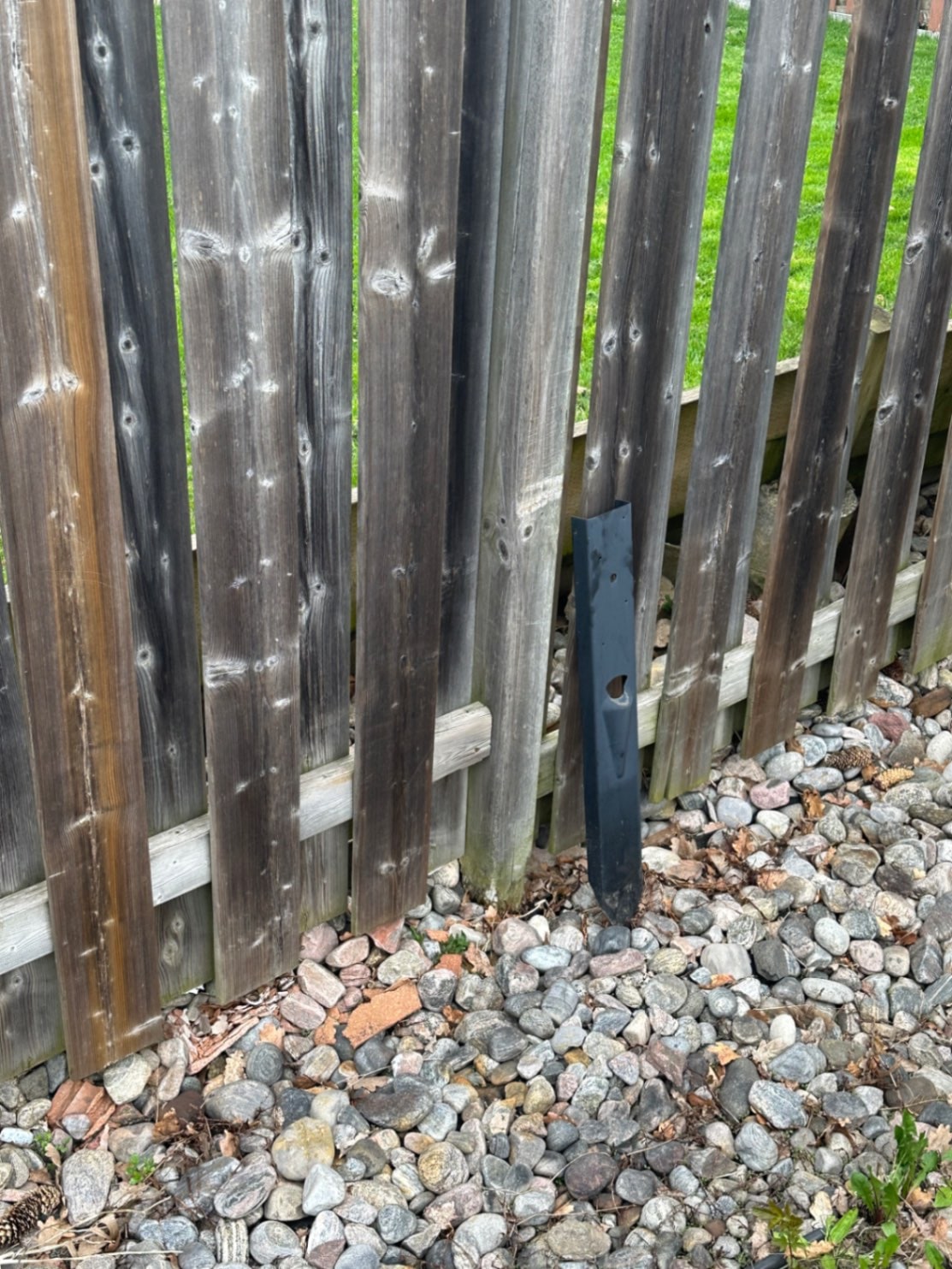 A loose black E-Z Mender leaning against the fence it used to hold, drive-in point and bolt holes visible — it worked free of the post and the soil — Georgina, Ontario, May 2026.