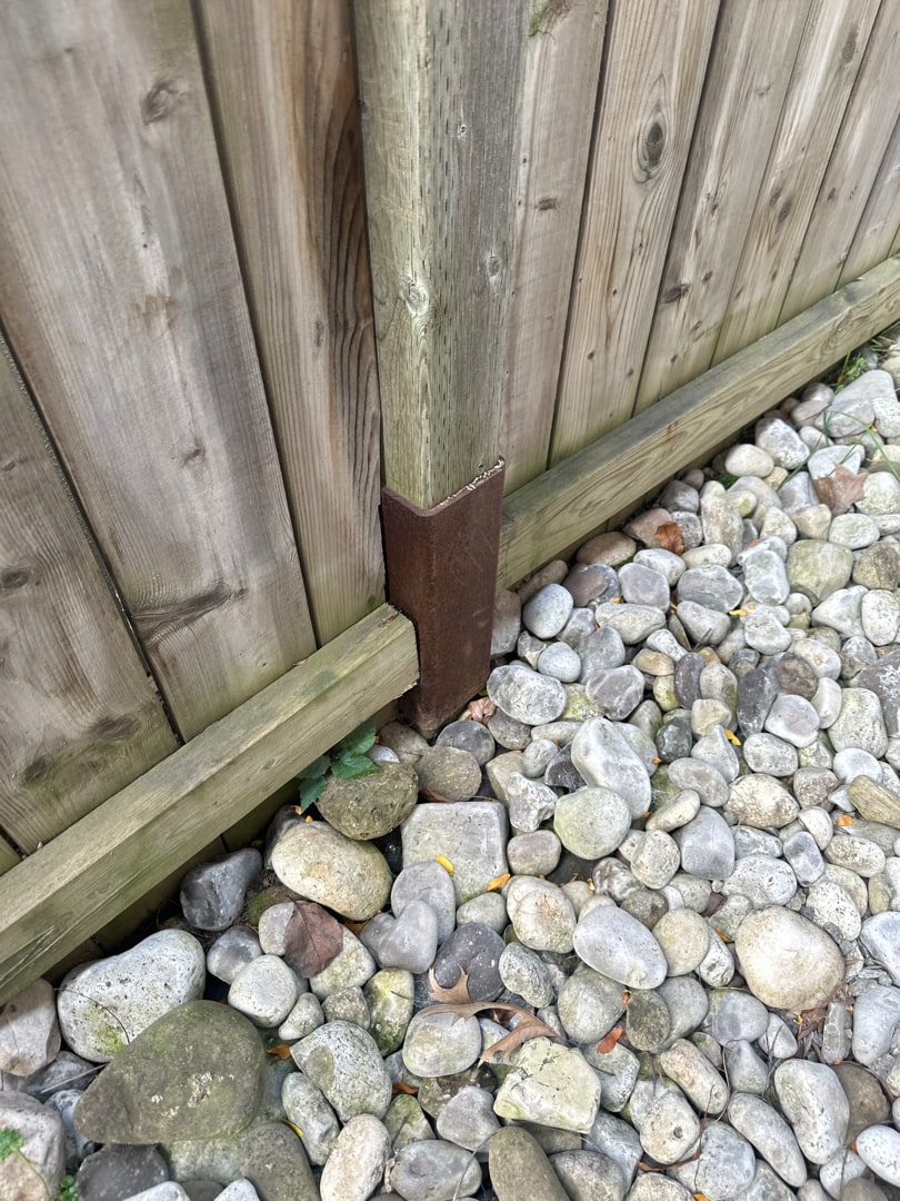 A mender plate rusted through end to end at a post base, the fence sagging onto its kickboard beside river rock — Niagara Falls, Ontario, September 2023.